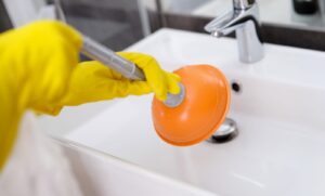 Plumber-with-rubber-gloves-cleaning-sink-with-plunger-in-bathroom